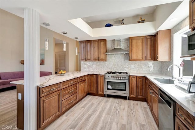 kitchen featuring wall chimney exhaust hood, light wood-style flooring, brown cabinets, stainless steel appliances, and a sink