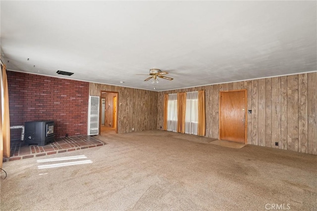 unfurnished living room with visible vents, a ceiling fan, a wood stove, carpet flooring, and wood walls