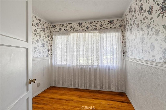 unfurnished room with a wainscoted wall, crown molding, wood finished floors, and wallpapered walls