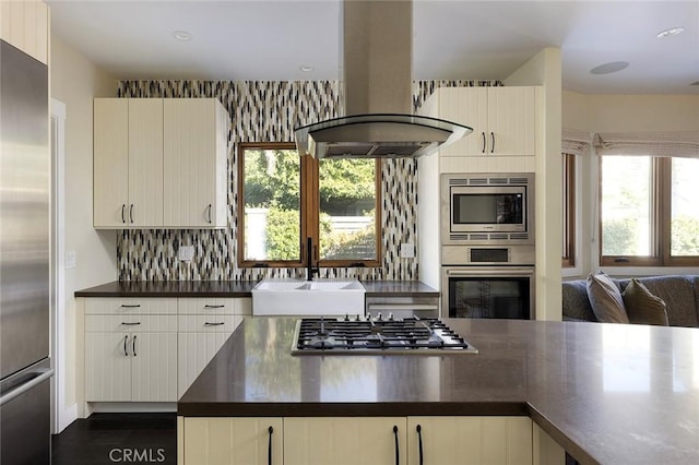 kitchen with island exhaust hood, stainless steel appliances, dark countertops, tasteful backsplash, and a sink