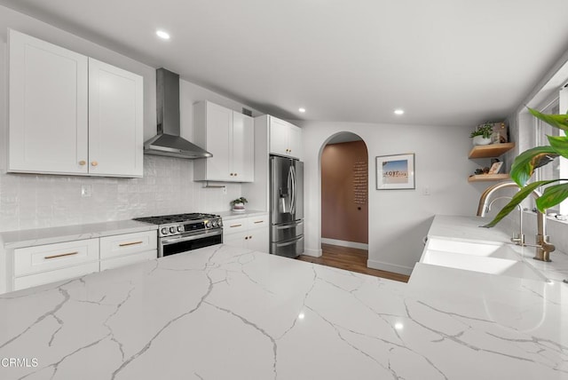 kitchen featuring arched walkways, wall chimney exhaust hood, appliances with stainless steel finishes, open shelves, and a sink