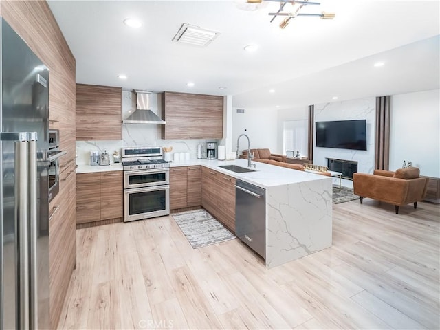 kitchen featuring brown cabinets, stainless steel appliances, a sink, wall chimney range hood, and modern cabinets