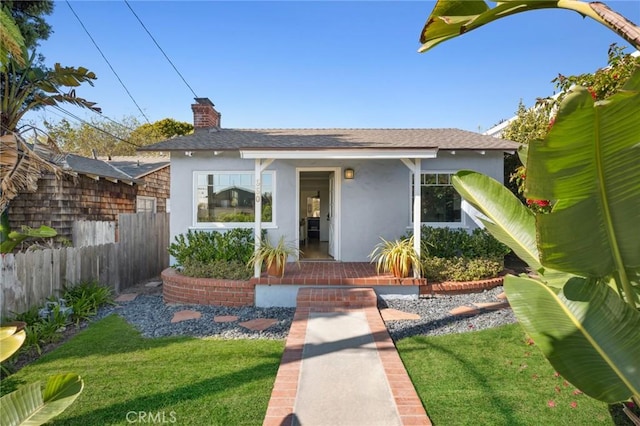 bungalow with a shingled roof, stucco siding, a chimney, fence, and a front yard