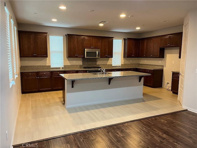 kitchen with stainless steel appliances, a sink, light wood finished floors, a kitchen bar, and a center island with sink