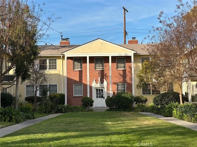 neoclassical / greek revival house with brick siding, a chimney, a front lawn, and stucco siding