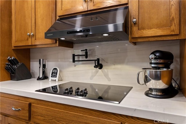 kitchen with black electric stovetop, light countertops, decorative backsplash, brown cabinetry, and under cabinet range hood