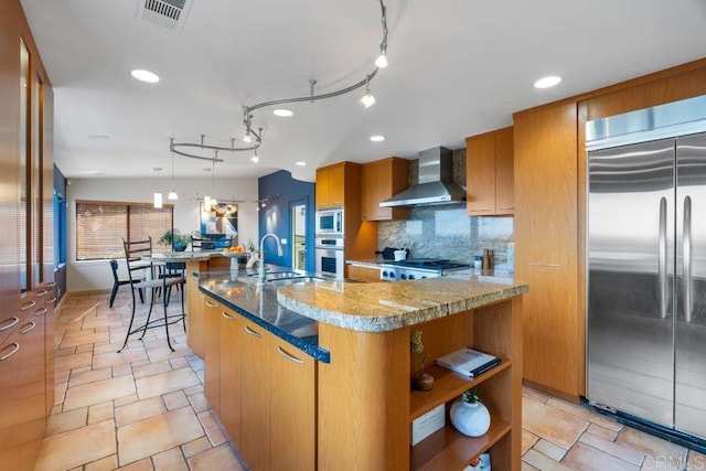 kitchen with visible vents, brown cabinetry, a sink, built in appliances, and wall chimney exhaust hood