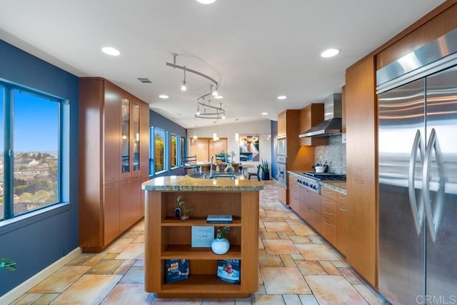 kitchen featuring stainless steel appliances, wall chimney range hood, backsplash, brown cabinets, and decorative light fixtures