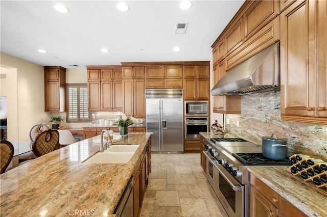 kitchen featuring a sink, light stone counters, stone tile floors, wall chimney exhaust hood, and built in appliances