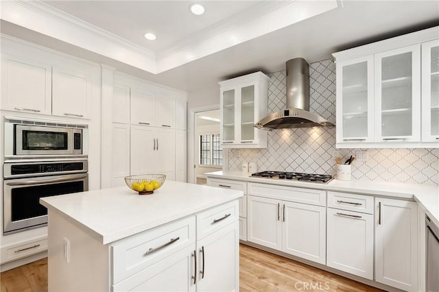 kitchen with wall chimney exhaust hood, appliances with stainless steel finishes, ornamental molding, a tray ceiling, and backsplash