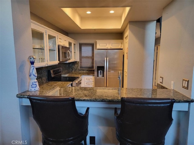 kitchen with a raised ceiling, glass insert cabinets, appliances with stainless steel finishes, a peninsula, and built in desk