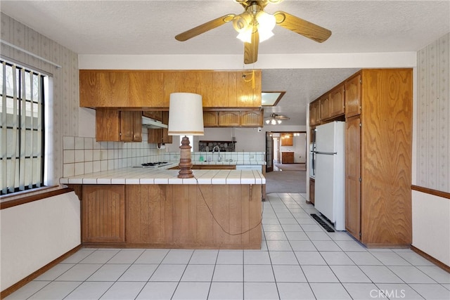 kitchen with tile countertops, a sink, white appliances, a peninsula, and wallpapered walls