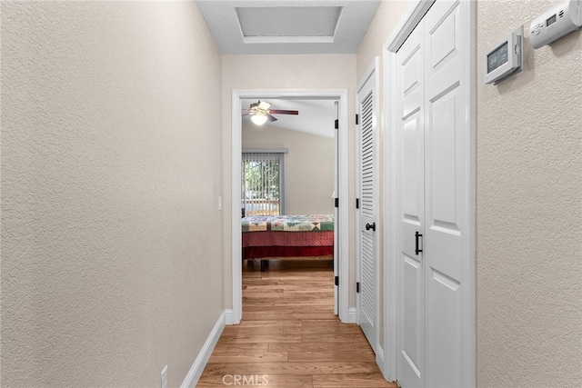 hallway with a textured wall, wood finished floors, attic access, and baseboards