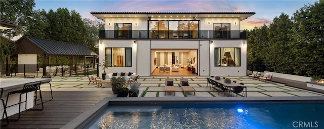 back of property at dusk with stucco siding, outdoor lounge area, a balcony, and an outdoor pool