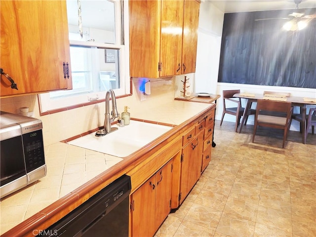 kitchen with tile countertops, stainless steel microwave, brown cabinetry, a sink, and dishwasher