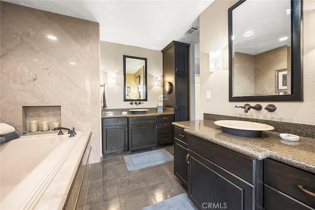 full bath featuring tile walls, visible vents, two vanities, a sink, and a bath