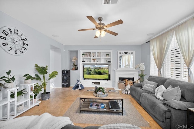 living room featuring a warm lit fireplace, ceiling fan, built in shelves, and tile patterned floors