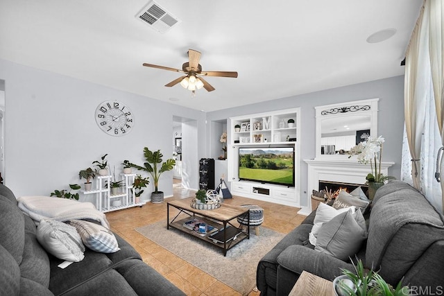 living room featuring a warm lit fireplace, ceiling fan, built in shelves, visible vents, and tile patterned floors