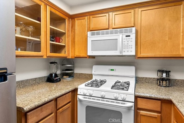 kitchen with white appliances, brown cabinetry, glass insert cabinets, and light stone countertops