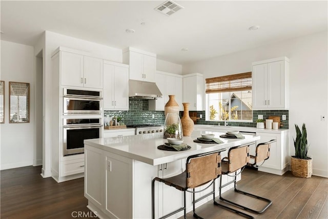 kitchen with light countertops, a kitchen island, double oven, under cabinet range hood, and visible vents