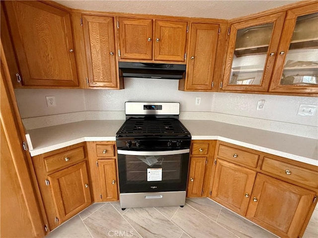 kitchen with light countertops, brown cabinetry, glass insert cabinets, under cabinet range hood, and stainless steel gas range oven