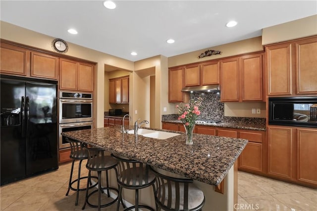 kitchen with a sink, black appliances, brown cabinetry, dark stone countertops, and a kitchen bar