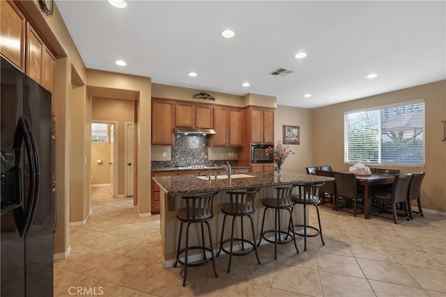 kitchen with tasteful backsplash, brown cabinets, a kitchen breakfast bar, under cabinet range hood, and black appliances