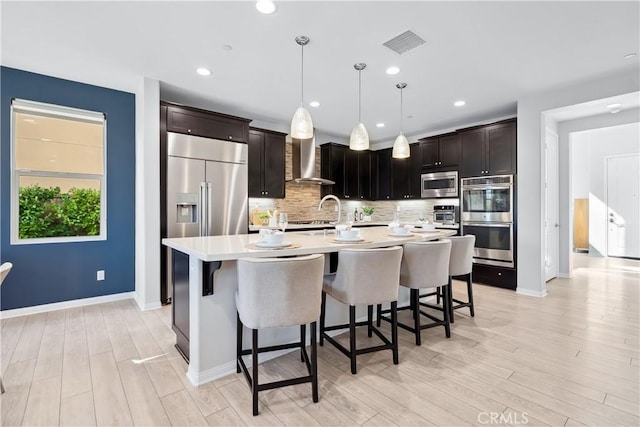 kitchen with a breakfast bar, tasteful backsplash, light countertops, built in appliances, and wall chimney exhaust hood