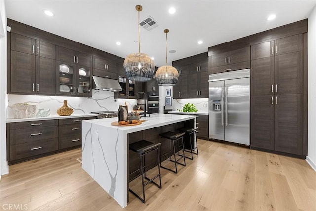 kitchen with a breakfast bar, light countertops, light wood-style floors, stainless steel built in refrigerator, and under cabinet range hood