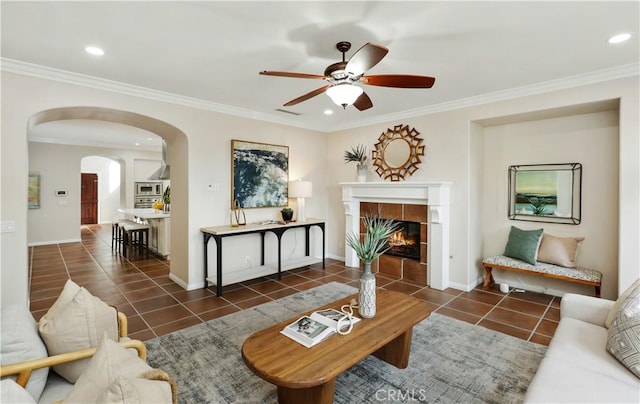 tiled living room with ceiling fan, arched walkways, ornamental molding, a tile fireplace, and recessed lighting