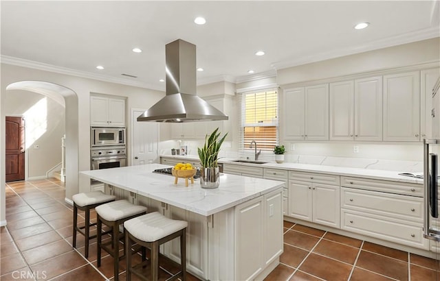 kitchen featuring arched walkways, white cabinets, island range hood, stainless steel appliances, and a sink