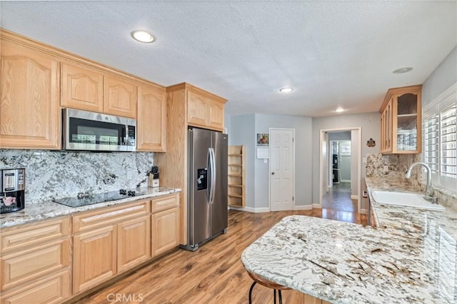 kitchen featuring light stone counters, glass insert cabinets, stainless steel appliances, light brown cabinetry, and a sink