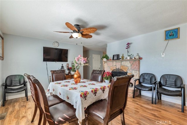 dining room with a stone fireplace, light wood-type flooring, a ceiling fan, and baseboards