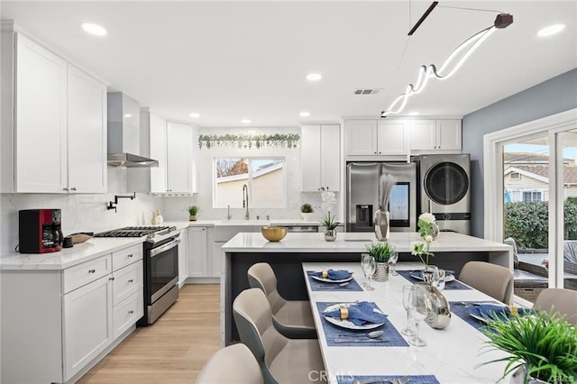 kitchen featuring white cabinets, appliances with stainless steel finishes, stacked washing maching and dryer, backsplash, and wall chimney exhaust hood