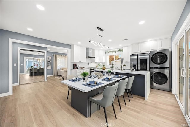 kitchen featuring wall chimney exhaust hood, a large island, appliances with stainless steel finishes, stacked washer / drying machine, and white cabinetry