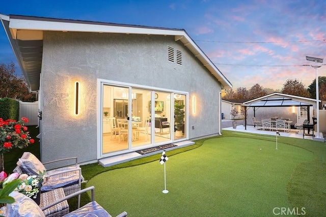 back of house at dusk with stucco siding, fence, a patio, and a gazebo