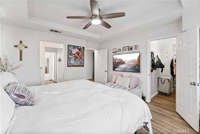 bedroom with a tray ceiling, light wood finished floors, visible vents, a spacious closet, and a ceiling fan