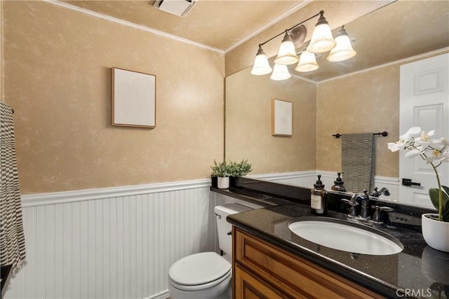 half bathroom featuring toilet, a wainscoted wall, vanity, visible vents, and ornamental molding