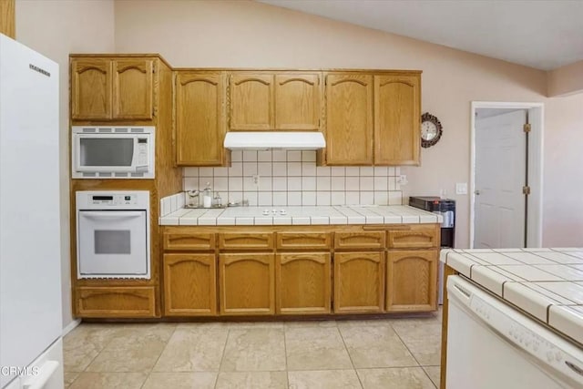 kitchen with tasteful backsplash, tile counters, vaulted ceiling, white appliances, and under cabinet range hood