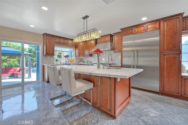 kitchen featuring tasteful backsplash, an island with sink, decorative light fixtures, stainless steel built in refrigerator, and under cabinet range hood