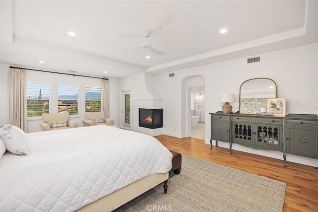 bedroom with wood finished floors, visible vents, a tray ceiling, a multi sided fireplace, and arched walkways