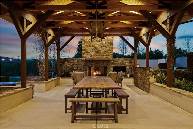 patio terrace at dusk with an outdoor stone fireplace, outdoor dining area, and a gazebo