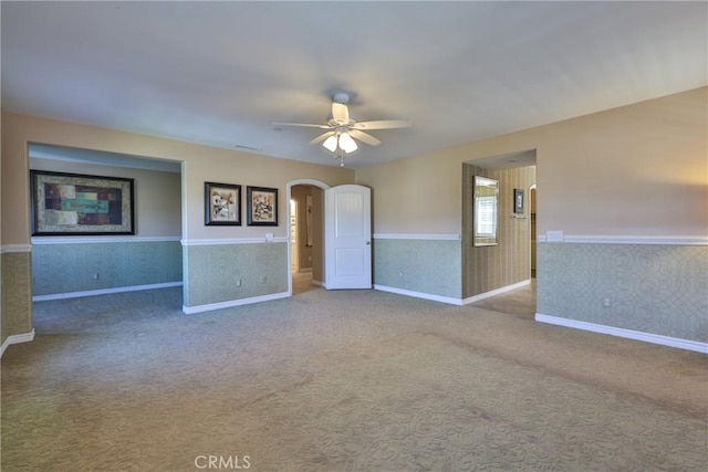 carpeted spare room featuring arched walkways, ceiling fan, baseboards, wainscoting, and wallpapered walls