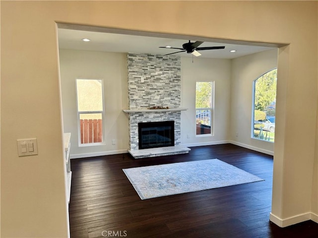 unfurnished living room featuring a stone fireplace, a ceiling fan, baseboards, and dark wood-style flooring