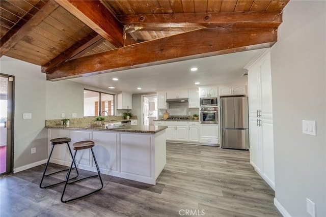 kitchen with appliances with stainless steel finishes, white cabinets, dark stone countertops, a peninsula, and under cabinet range hood