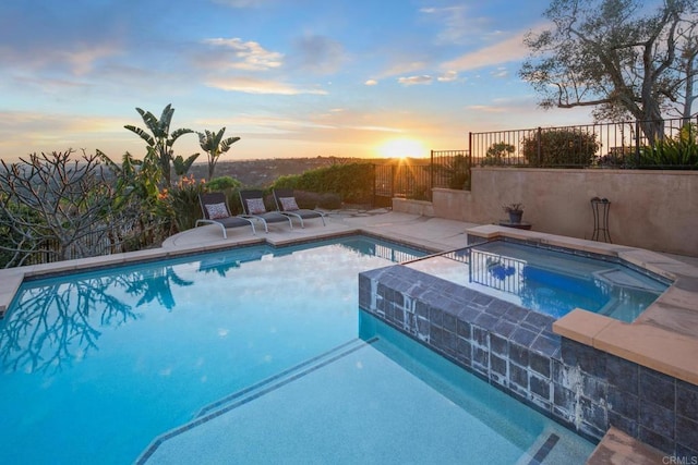 pool at dusk featuring an outdoor pool, fence, and an in ground hot tub