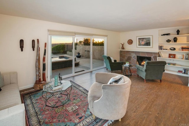 living room featuring built in shelves, a stone fireplace, baseboards, and wood finished floors