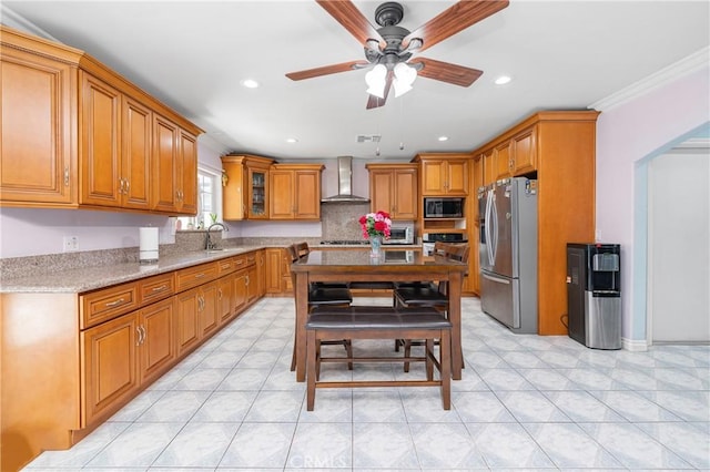 kitchen featuring brown cabinetry, light stone counters, stainless steel appliances, wall chimney range hood, and a sink