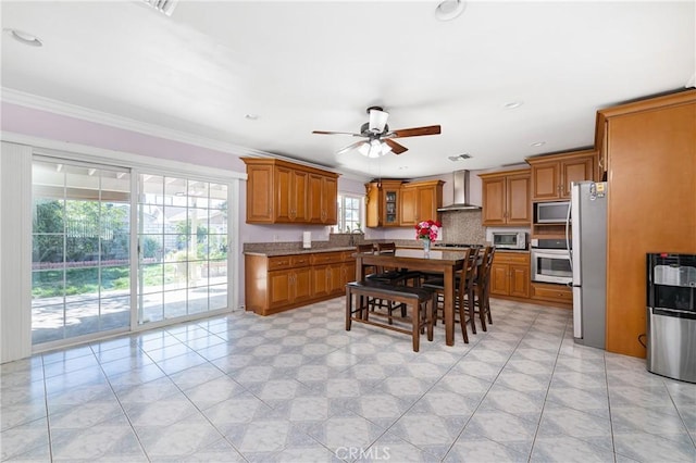 kitchen with ceiling fan, wall chimney exhaust hood, appliances with stainless steel finishes, brown cabinets, and ornamental molding