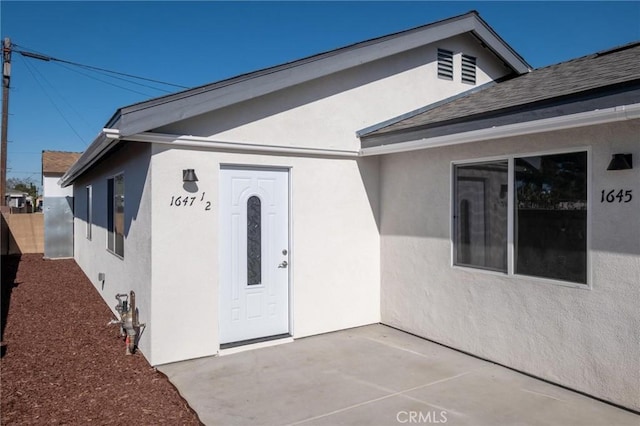 entrance to property with roof with shingles, a patio area, fence, and stucco siding